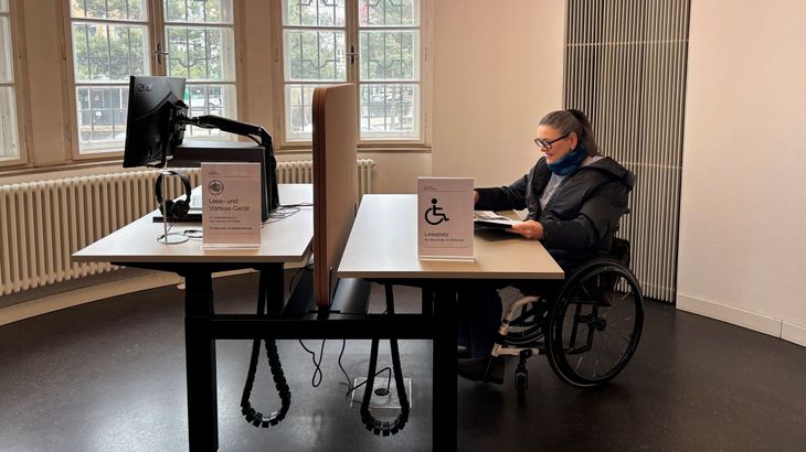Frau im Rollstuhl liest ein Buch am höhenverstellbaren Arbeitsplatz, gegenüber vom Bildschirmlesegerät mit Vorlesefunktion. 